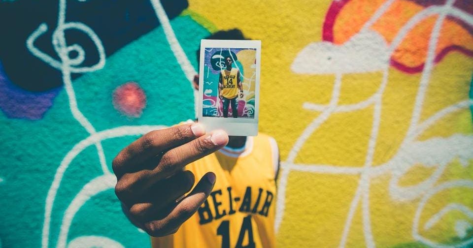 Person holding up a polaroid photo of themselves against a colorful mural