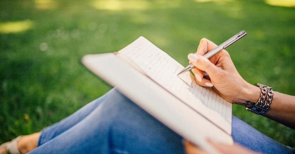 Person sitting on the grass writing in a notebook with a silver pen